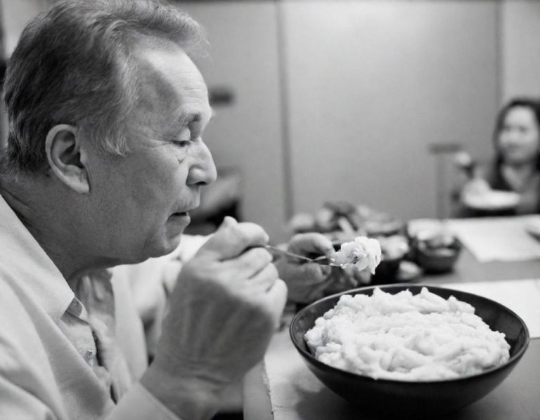 Homme âgé savourant un grand bol de purée, assis à une table avec une femme en arrière-plan.