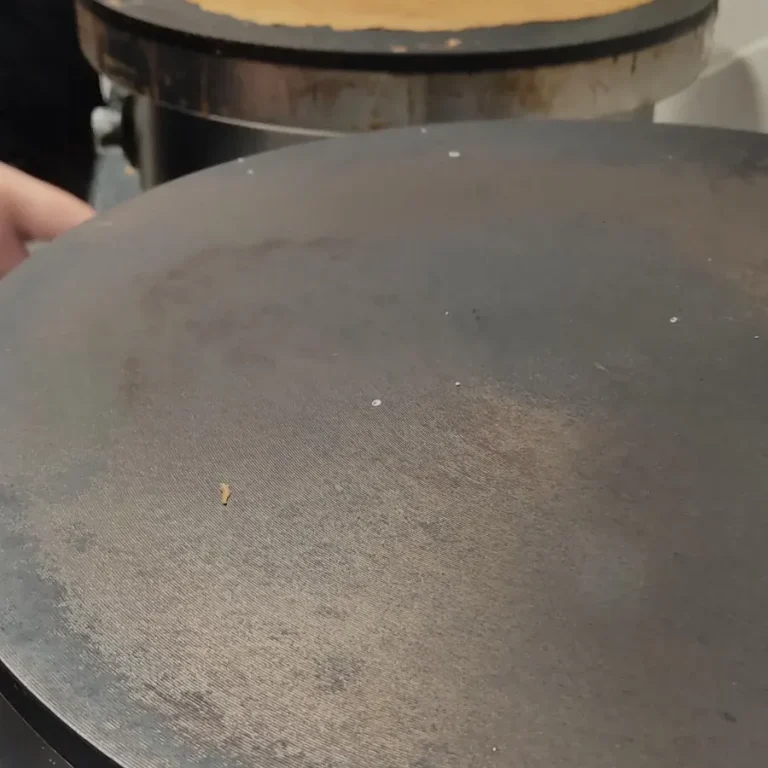 Une crêpe dorée cuite sur une crêpière, avec des mains en arrière-plan.